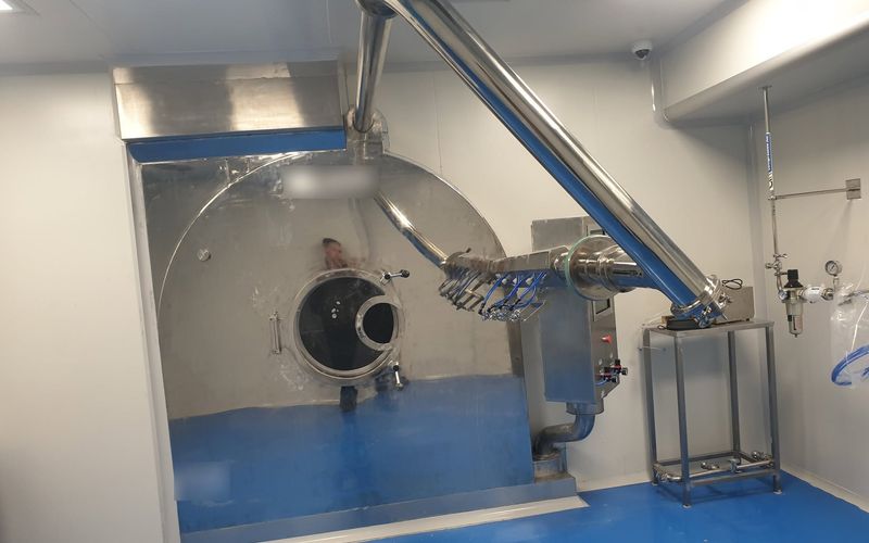 Stainless-steel tablet coating drum with spray-gun arm and exhaust duct in a pharmaceutical cleanroom